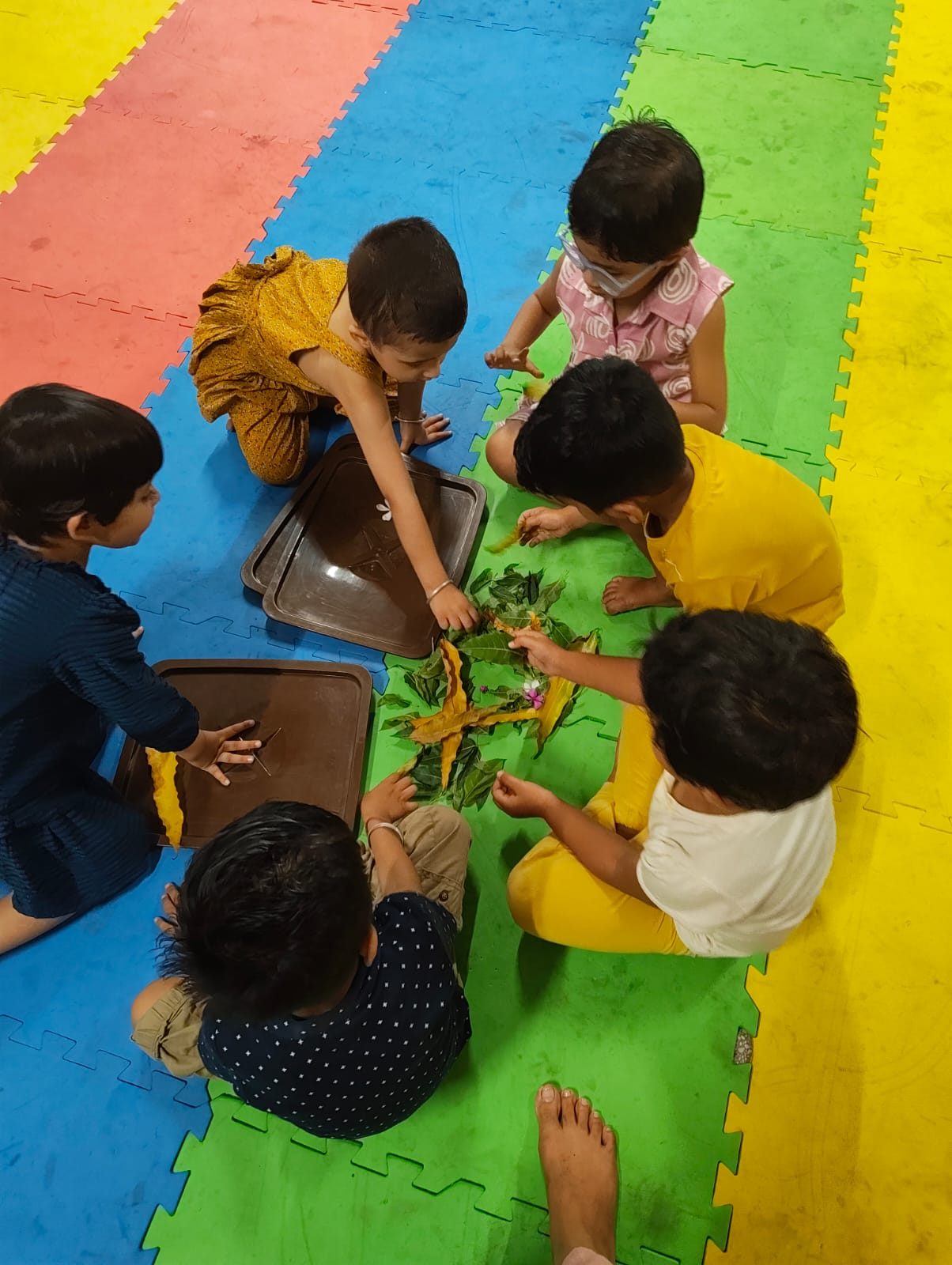 Six children kneeling on the coloured mat, arranging leaves they have collected on brown trays.